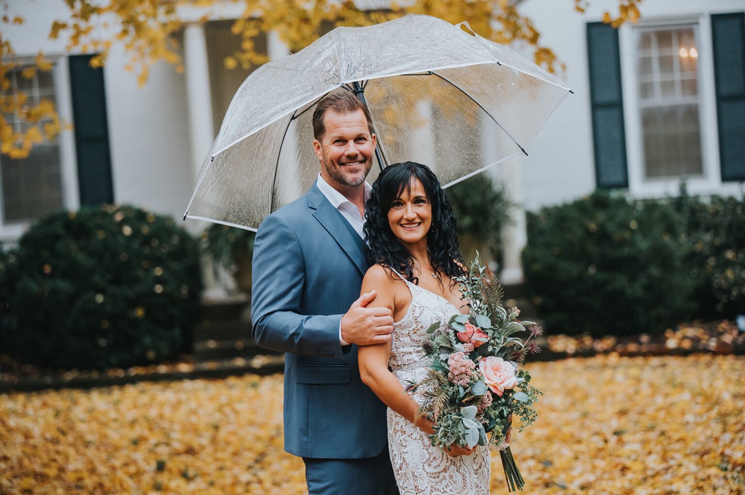 Elopement Wedding A couple stands closely under a transparent umbrella on a rainy day. The man, in a light blue suit, smiles as he embraces the woman. The woman, dressed in a white lace gown, holds a bouquet of flowers. They're surrounded by autumn leaves in front of a white house with green shutters. Elopements Inc